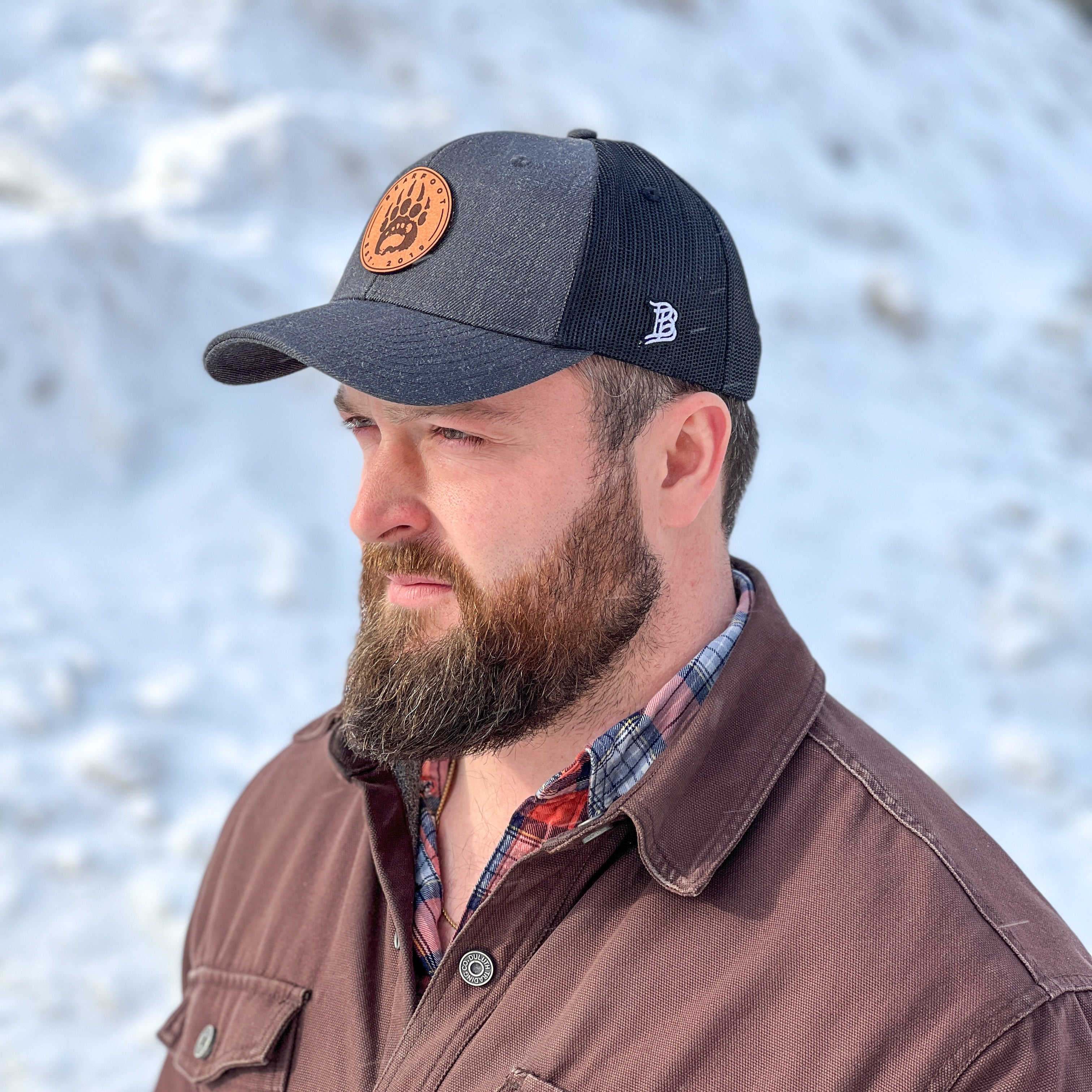 A black and gray Bearfoot Leather Patch Trucker Hat with a circular orange logo featuring a black bear paw print in the center.